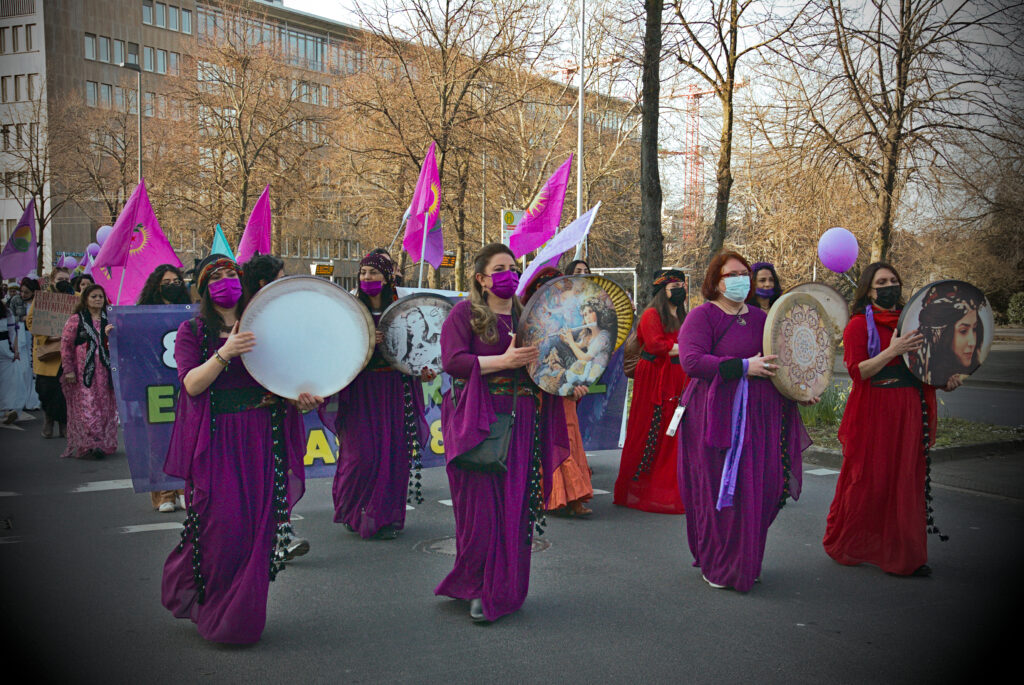An der Spitze der feministischen Demonstration laufen lila und rot gekleidete weiblich gelesene Personen, die auf Trommeln spielen. Im Hintergrund sind lila Fahnen zu sehen.
