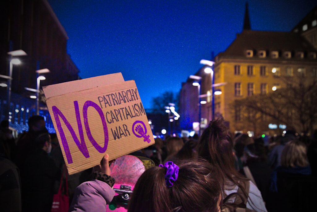 Bochum bei Nacht. Eine weiblich gelesene Person hält ein Schild aus der Demonstration auf dem steht: "No Patriachy, no Capitalism, no War"